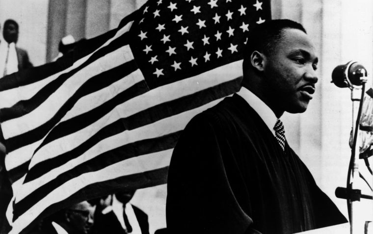 A black and white picture of Dr Martin Luther King Jr. speaking with the USA flag in the background.