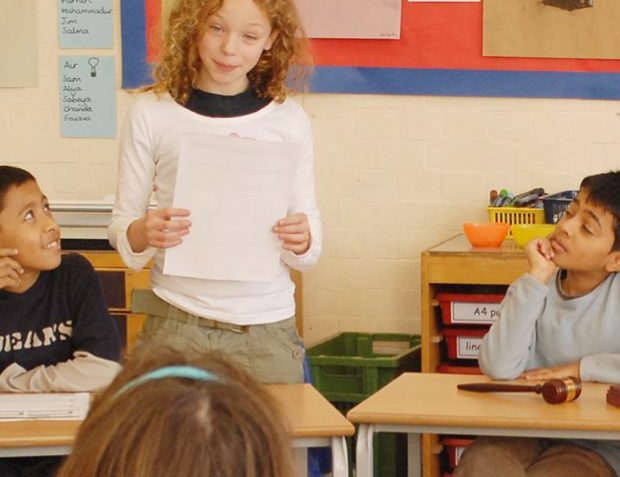 A primary school standing inside a classroom and holding a piece of paper in her hand while speaking to the pupils sitting down.