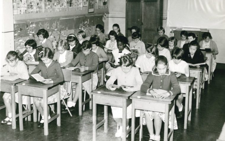 Black and white photo of a classroom of children sitting at old wooden desks.