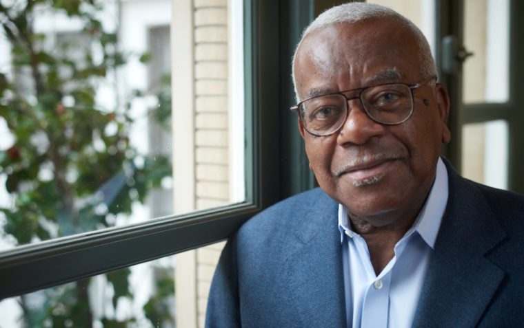 Sir Trevor McDonald close up, next to a window with trees behind