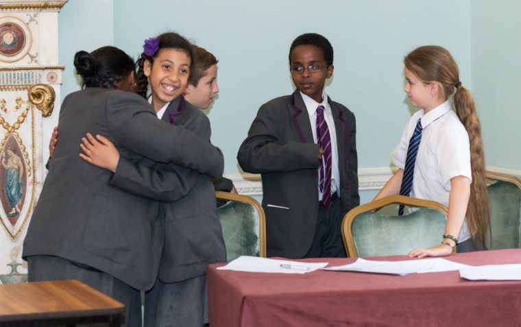 Five primary school pupils (two boys and three girls) inside a classroom. Two girls are hugging and the others are having a conversation.