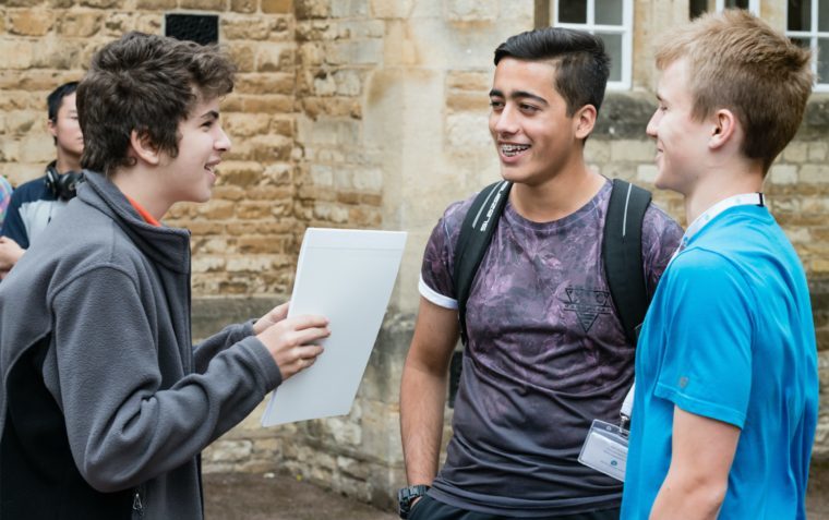 Three young male students of the Debate Academy summer school standing outside the building talking and laughing. One is holding a piece of paper in his hand and the other has a