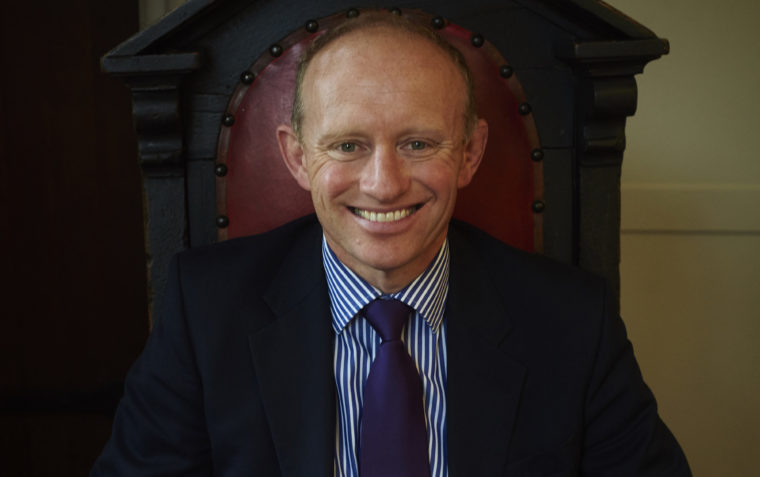 A photograph of Mark Mortimer sitting on headmaster's chair at school