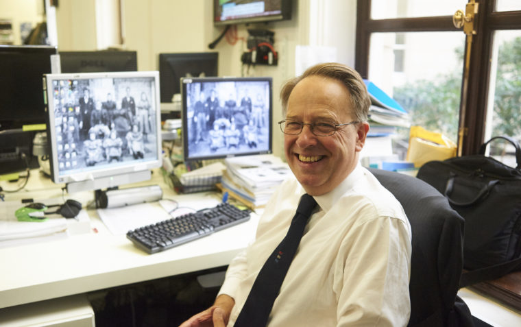 A picture of smiling Michael Crick wearing contact lens glasses sitting inside his office with two computer screens and a keyboard on the desk.