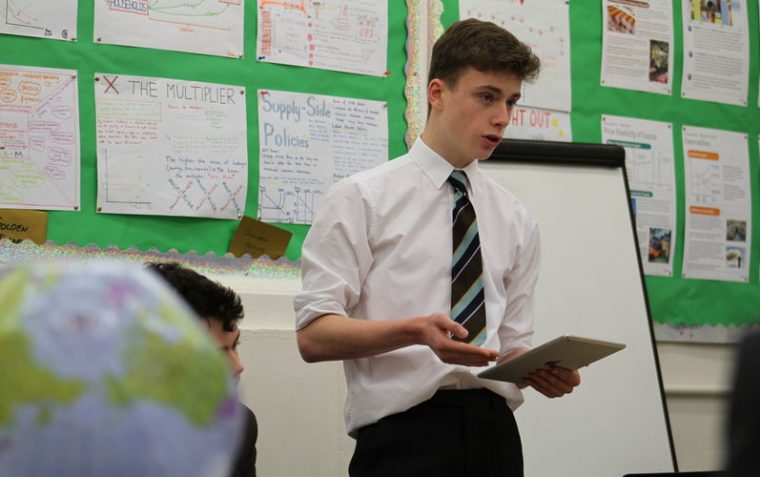 A male secondary school student standing in the classroom while reading from the notepad in his hand.