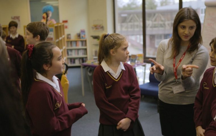 A teacher explaining a concept while 3 of her students listen to her carefully. There are more pupils a bit far away in the background.