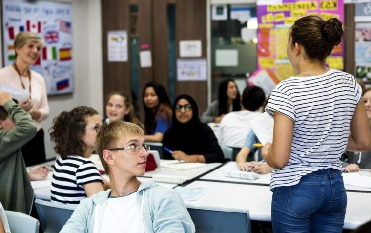 A female Secondary School Teacher holding books in her hand in the classroom with students sitting down and one female standing.