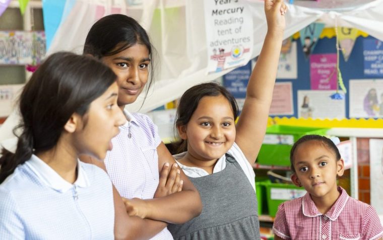 Four primary school pupils (all girls) standing inside a classroom, and one raised her hand in the air.