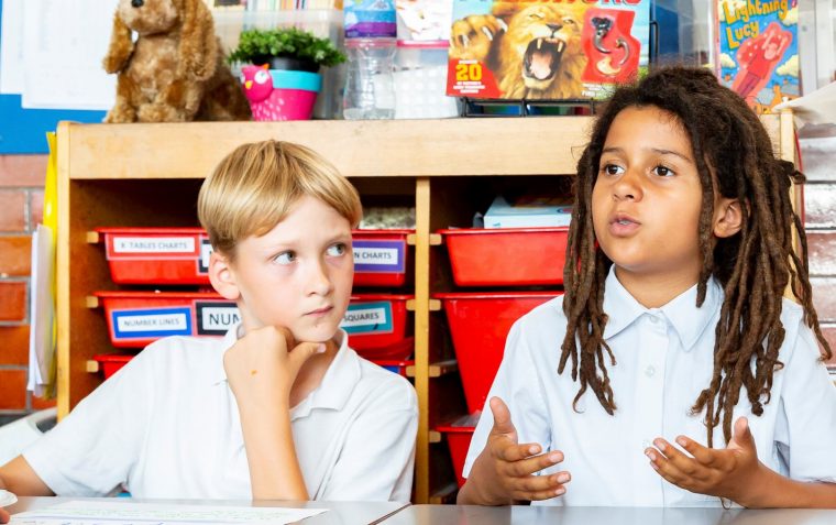 Two primary school students. The one speaking and making hands gesture. The other listening carefully and watching with left hand under his chin whiles looking at his classmate.
