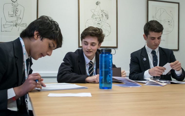 Three secondary school sitting around a table inside a classroom. One is reading from a book and the other is folding a paper.