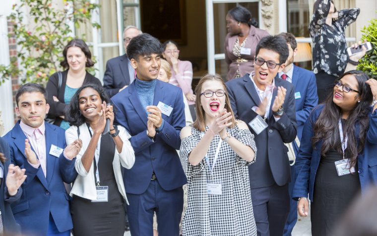 A group of secondary school students standing outside a building while clapping and cheering.