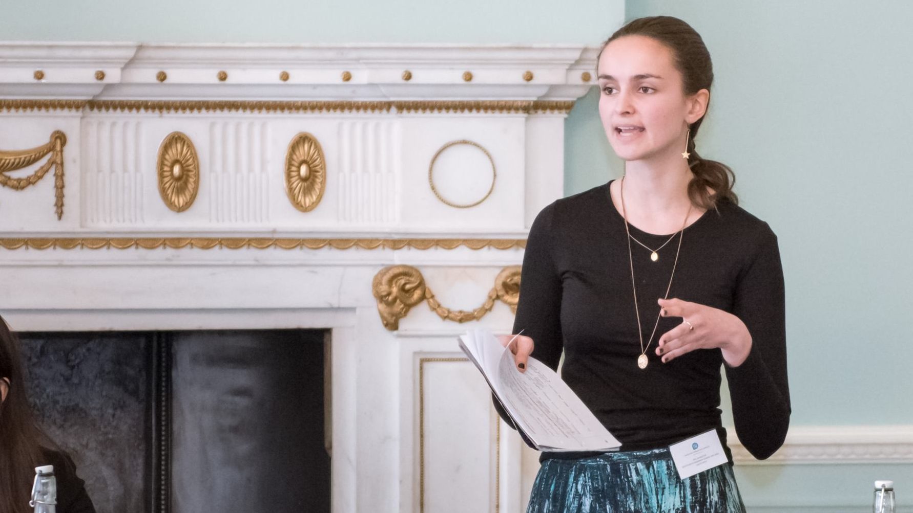 A female tutor with a notepad on her hand is standing whiles speaking.
