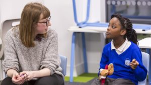 A female teacher and a girl pupil sitting down inside a classroom. The pupil is speaking to the teacher and holding a little teddy bear in her hand.