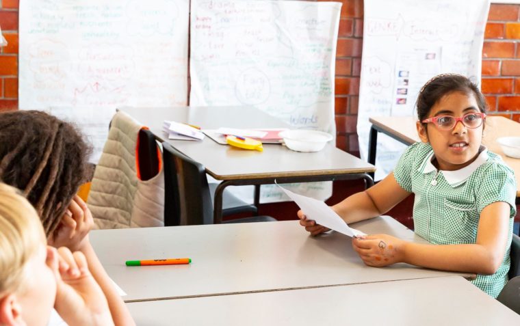 A clos-up of a young girl pupil sitting inside a classroom wearing contact lens glasses and holding a piece of paper.