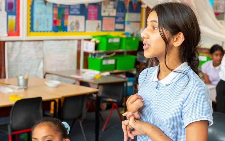 A clos-up of a young girl pupil standing and speaking inside a classroom with other pupils in the background.