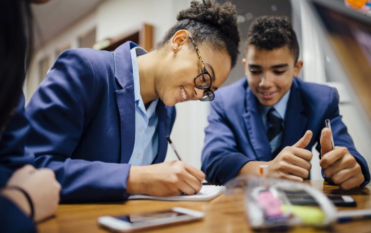 A male and a female secondary school students sitting down. The female is wearing lens glasses and writing on a notepad with heads bow and a smile. The male has both thumbs finger up and looking at her writing. There is a mobile phone and stica transparent plastic bag.