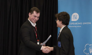 Louis Preston holding a piece of paper in his left hand and shaking a young student right hand on stage.