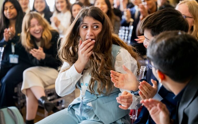 Young peoplp sat in a room with one young woman at the focus shocked with her hand in front of her mouth.
