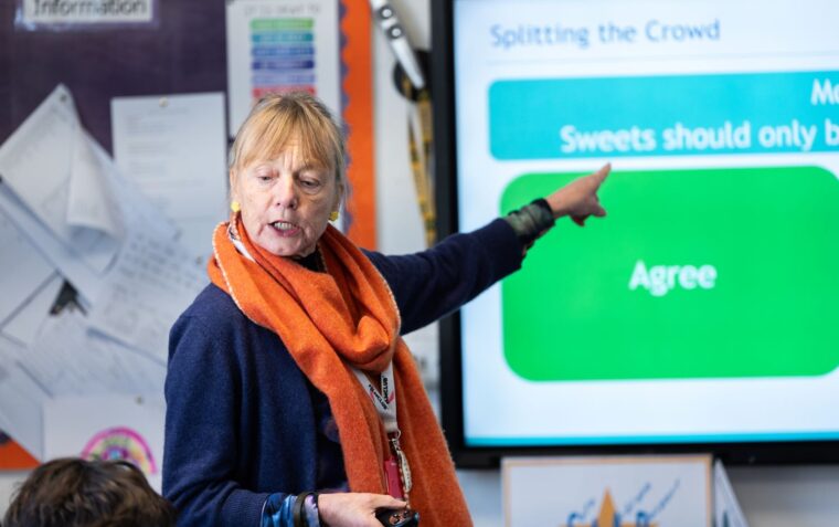 A female faciliator/teacher stood at the front of a classroom, pointing at an interactive screen facing the class in front of her